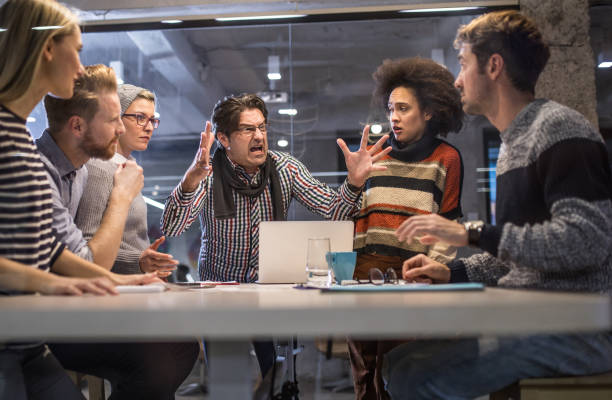 A group of people sitting around a table in a modern office setting, engaged in a tense discussion. A man in the center is angrily gesturing and shouting, while others display mixed reactions of discomfort, defensiveness, and frustration. The image represents conflict, misunderstandings, and interpersonal tensions, highlighting reasons why people might dislike or clash with others.